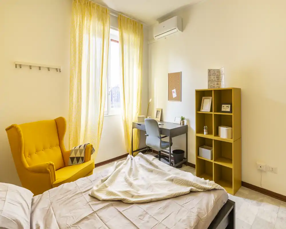 Camera da letto luminosa e accogliente con letto in primo piano, una poltrona gialla e una scrivania ordinata vicino alla finestra — arredamento caldo e luce naturale.