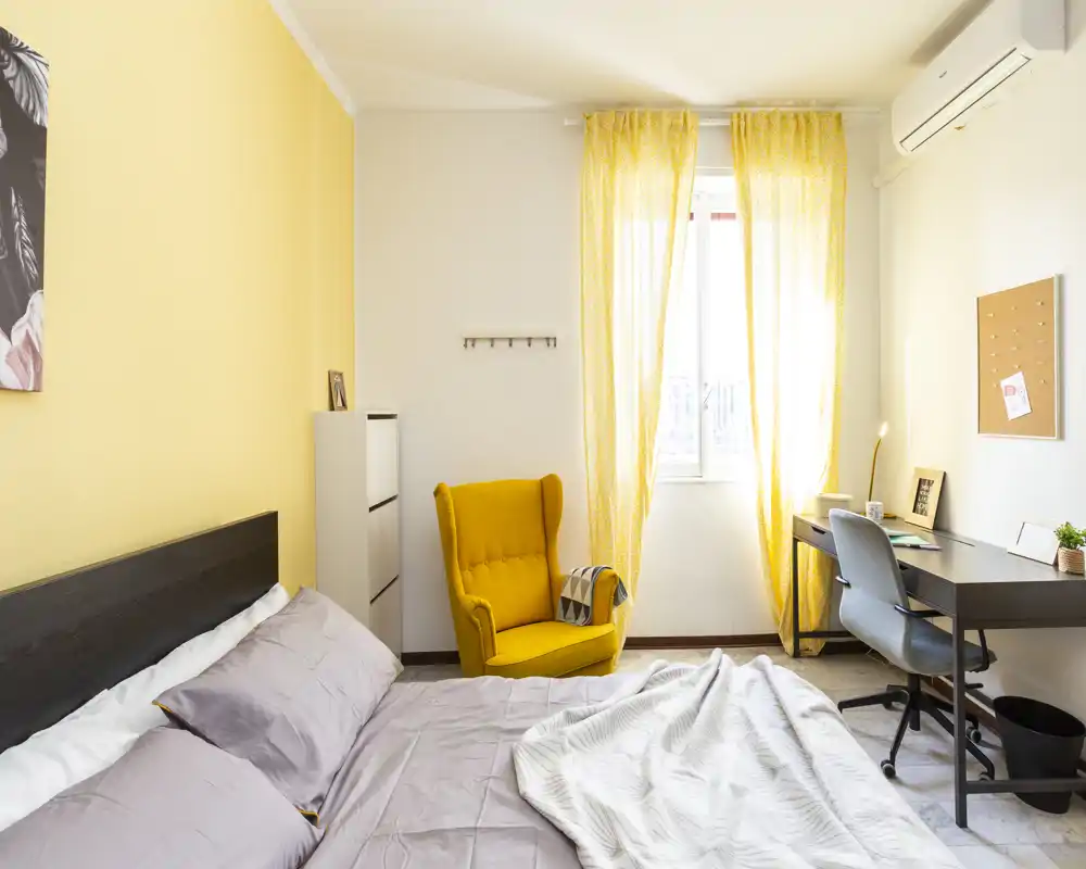 Camera da letto luminosa e ben curata con un comodo letto matrimoniale, una poltrona gialla accogliente e una scrivania vicino alla finestra che lascia entrare la luce naturale.
