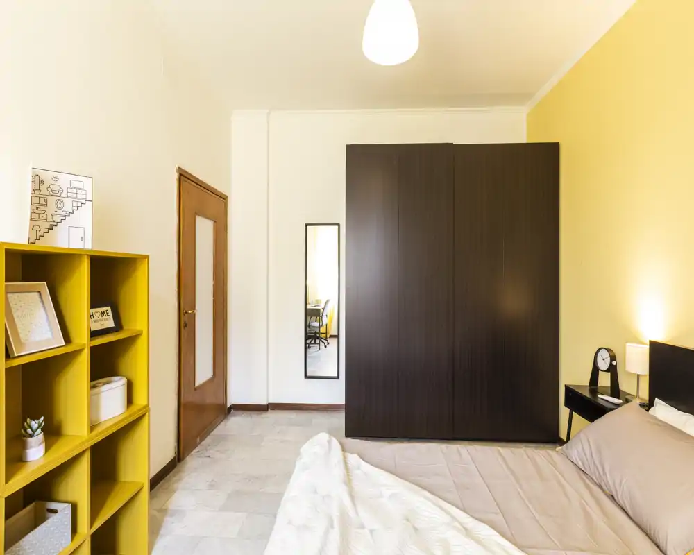 Camera da letto luminosa e ben arredata con letto matrimoniale, armadio scuro e una libreria gialla; illuminazione calda e arredo moderno e essenziale.