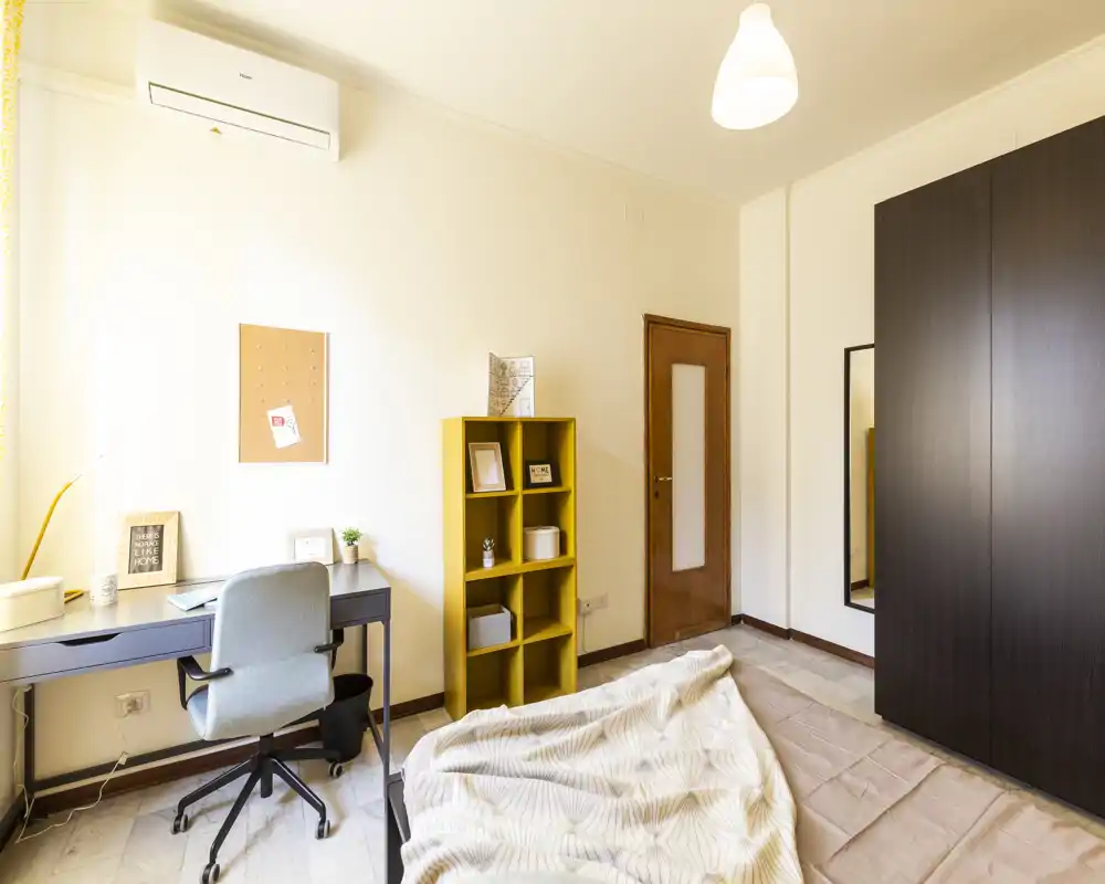 Camera da letto luminosa e ben arredata con letto visibile, angolo studio con scrivania e sedia, armadio e scaffale decorativo — perfetta per un annuncio in affitto che evidenzia comfort e funzionalità.