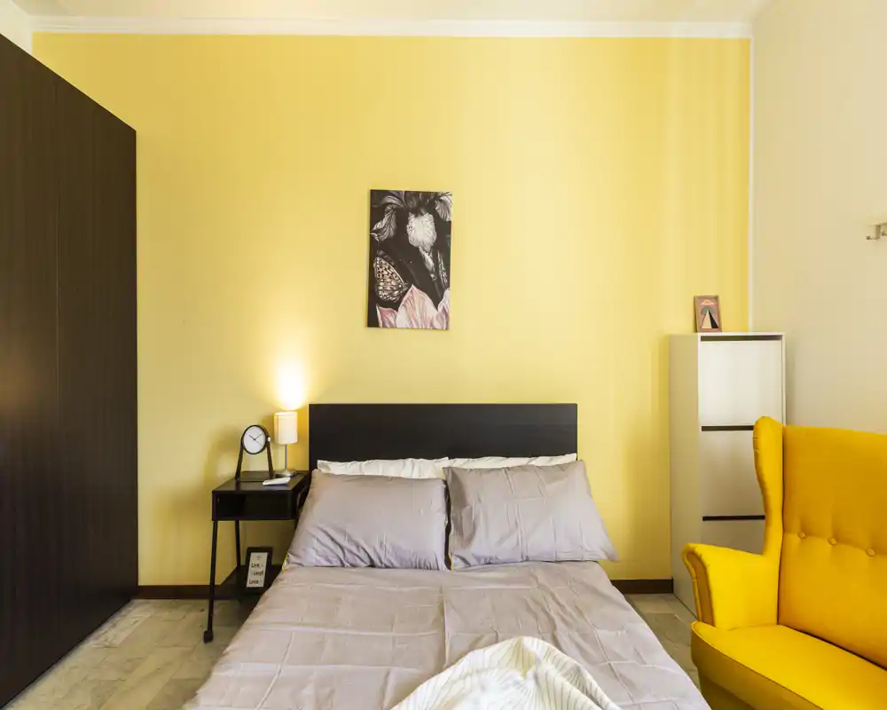 Camera da letto luminosa e ben allestita con letto matrimoniale, comodino con lampada, quadro sopra la testata e una poltrona gialla che crea un piacevole accento caldo.