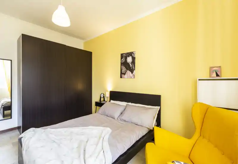 Camera da letto luminosa e accogliente con letto matrimoniale, comodino e una poltrona gialla vivace; biancheria neutra e parete calda per un’atmosfera contemporanea.
