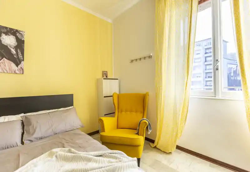 Camera da letto luminosa e accogliente con letto matrimoniale, biancheria morbida e una poltrona gialla vicino a una finestra che illumina la stanza con luce naturale.