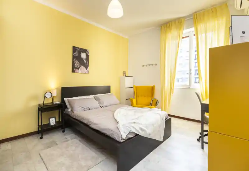 Camera da letto luminosa e accogliente con letto matrimoniale, comodino, poltrona gialla e luce naturale proveniente da una grande finestra.