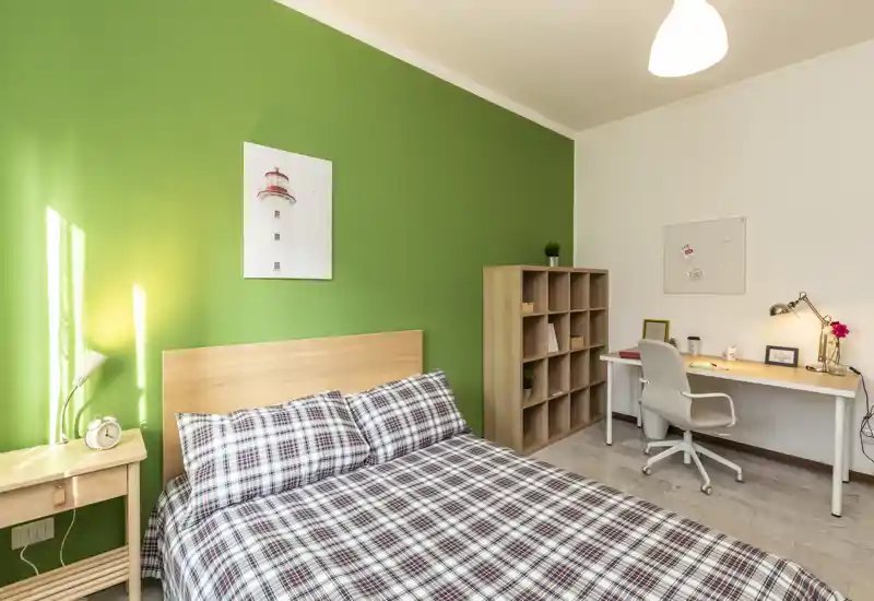 Bright, neatly staged bedroom with a double bed against a green accent wall, bedside table and a tidy study desk — ideal for comfortable short or long stays.