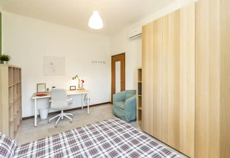 Well-lit bedroom with a plaid-covered bed in the foreground, a study desk and shelving along the wall, and a comfortable armchair. Neutral tones and wooden wardrobe create a calm, cozy atmosphere ideal for rest and work.
