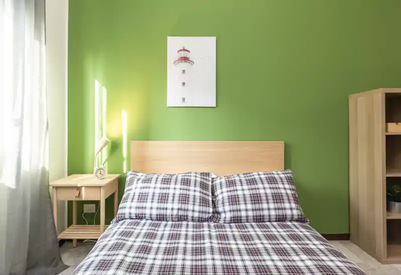 Bright, cozy bedroom with a central double bed dressed in patterned bedding, a wooden headboard, bedside table with lamp and a wardrobe on the right. Soft natural light from the window highlights the fresh green accent wall.