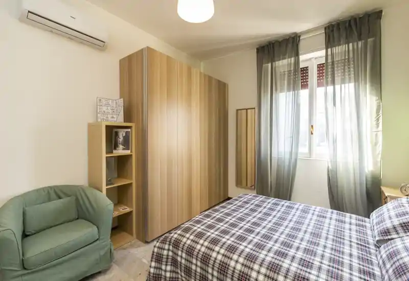 Bright, cozy bedroom with a double bed dressed in patterned linens, a wooden wardrobe, small armchair and natural light from a window.