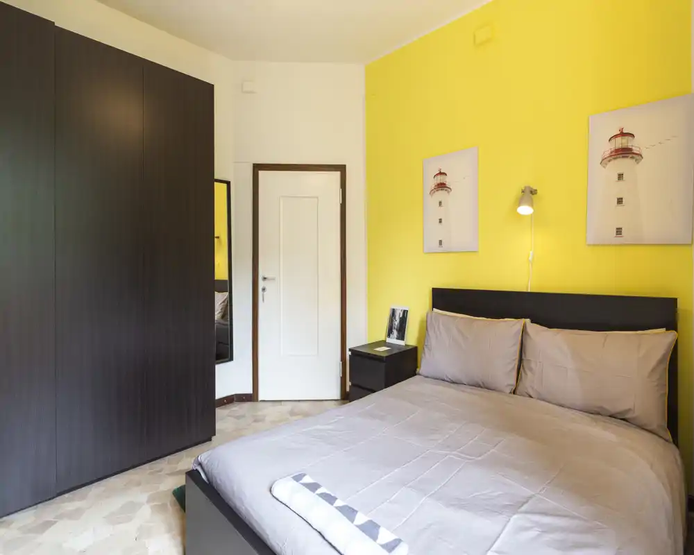 Bright, well-composed bedroom with a double bed, neutral linens and a bold yellow accent wall. The room features a large dark wardrobe, bedside table and simple wall art, ideal for rental listings highlighting a cozy, modern sleep space.