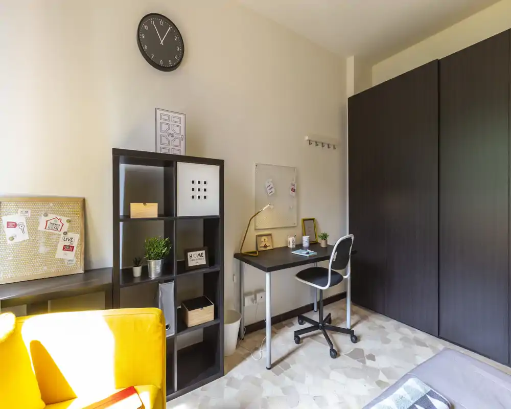 Bright, well-styled living area with a compact desk, shelving unit, wardrobe and a cozy yellow armchair — ideal for a single occupant or student.
