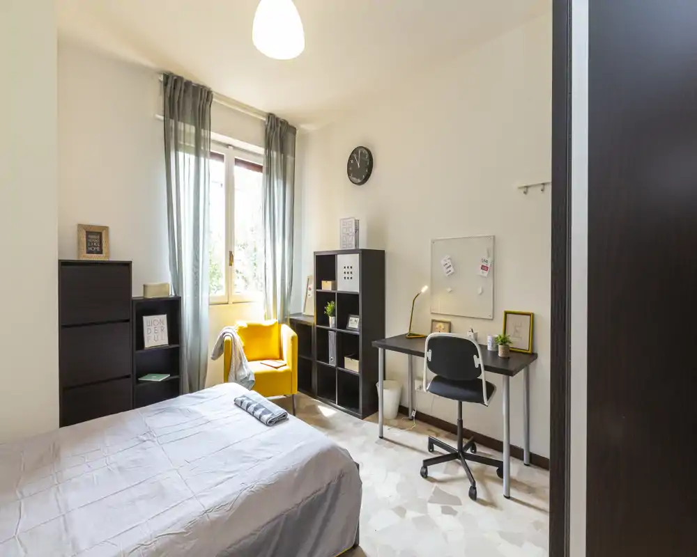 Bright, well-styled bedroom with a double bed in the foreground, a cozy yellow armchair by the window, shelving and a desk — ideal for both rest and study.