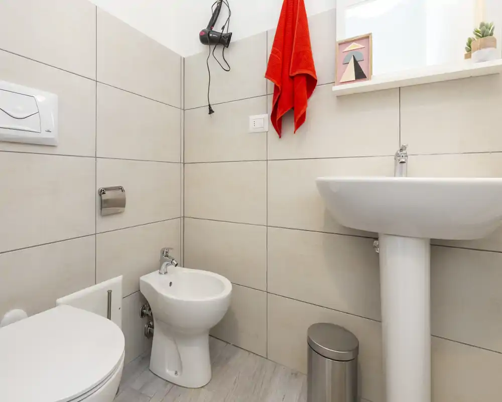 Bright, compact bathroom with a toilet, bidet and pedestal sink set against large neutral tiles; includes a red towel and small shelf decor.