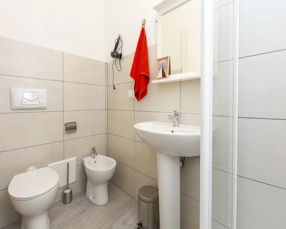 Compact, modern bathroom with toilet, bidet, pedestal sink and a shower enclosure. Neutral tiles and bright lighting create a clean, fresh atmosphere.