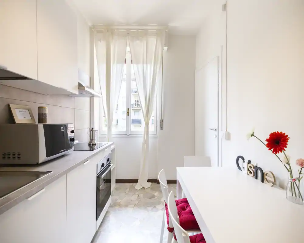 Bright modern kitchen with white cabinets, countertop appliances and a small dining table with red seat cushions — airy natural light from the window creates a fresh, welcoming atmosphere.