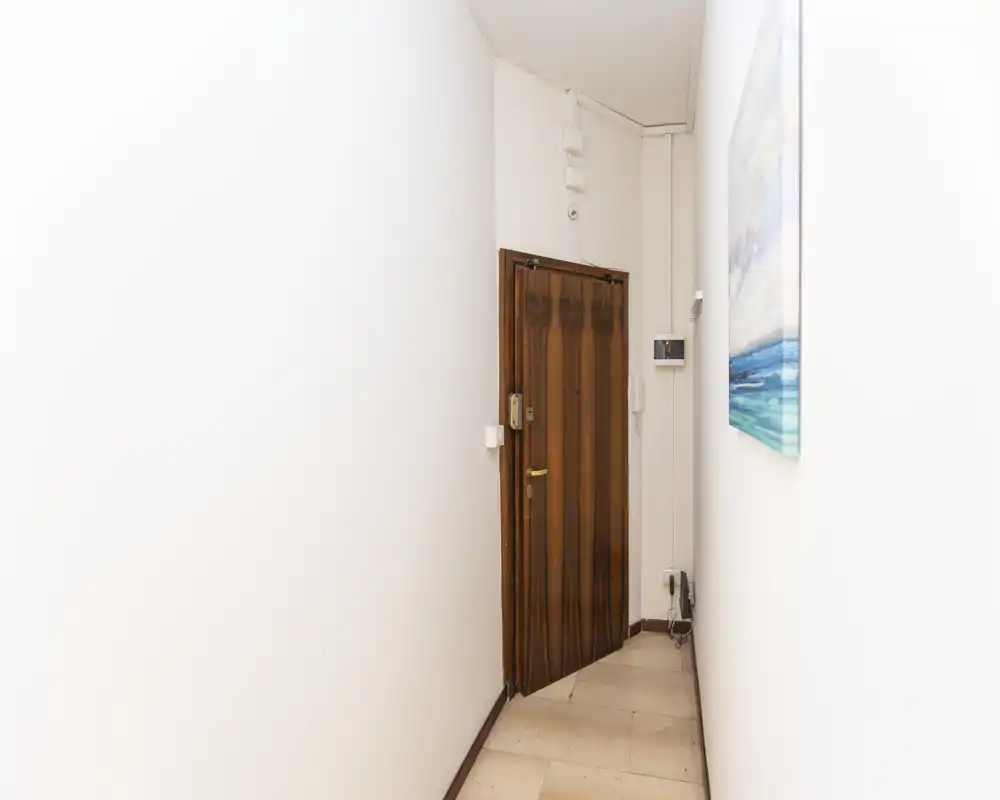 Narrow entrance corridor with a wooden front door, intercom and simple floor tiles; bright white walls create a clean, minimalist look.