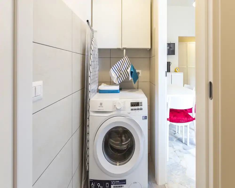 Compact laundry niche with a front-loading washing machine, wall cabinets above and an ironing board stored at the side; bright and tidy utility space connected to the dining area.