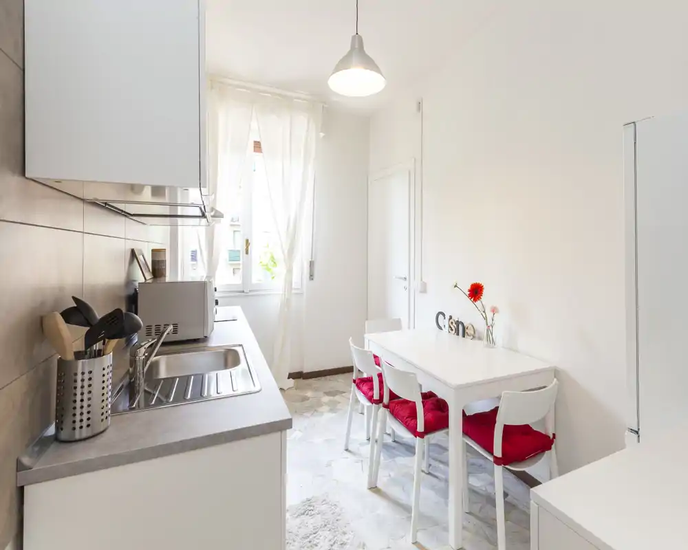 Bright compact kitchen with a counter sink, microwave and a small dining table with four chairs—white furnishings and red seat cushions give a fresh, cozy look.