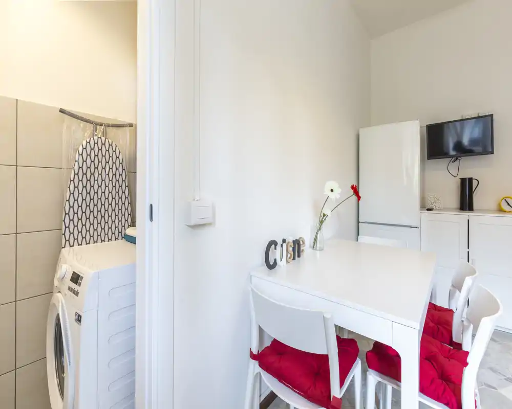 Compact modern kitchen corner with a white dining table, four chairs with red cushions, a tall fridge, and a small wall-mounted TV; bright, clean and pleasantly styled — ideal for a small apartment listing.