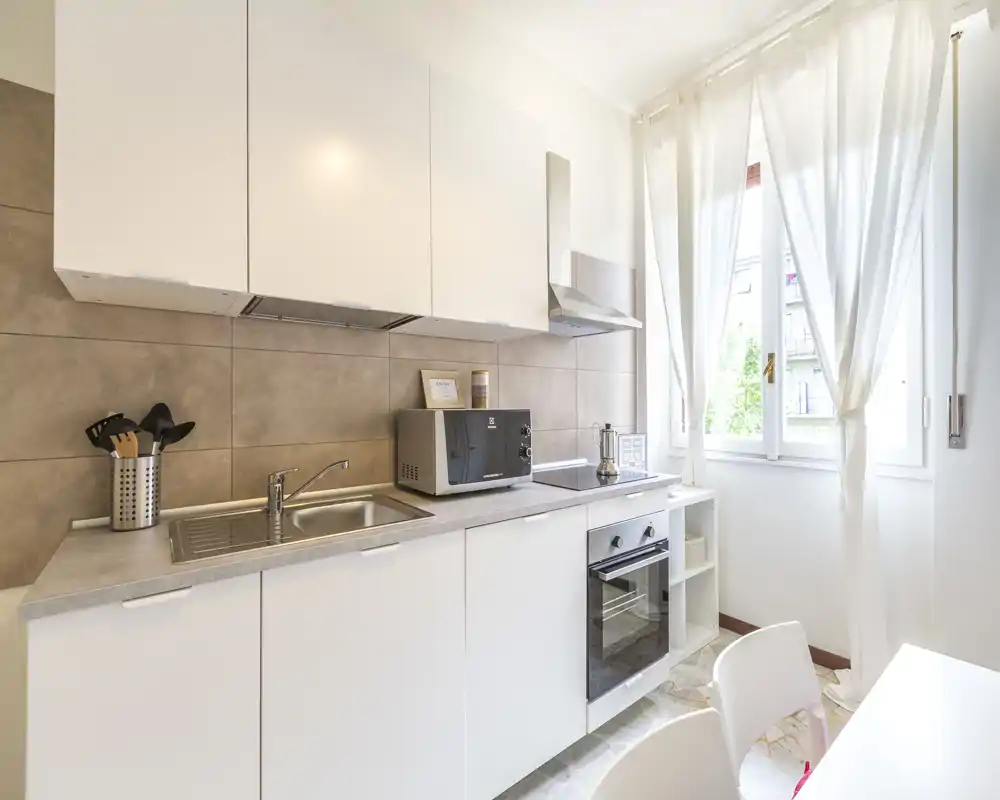 Bright, modern kitchenette with white cabinets, integrated oven, microwave and a sink under natural daylight. Minimal, clean styling and a small dining area make it ideal for compact city rentals.