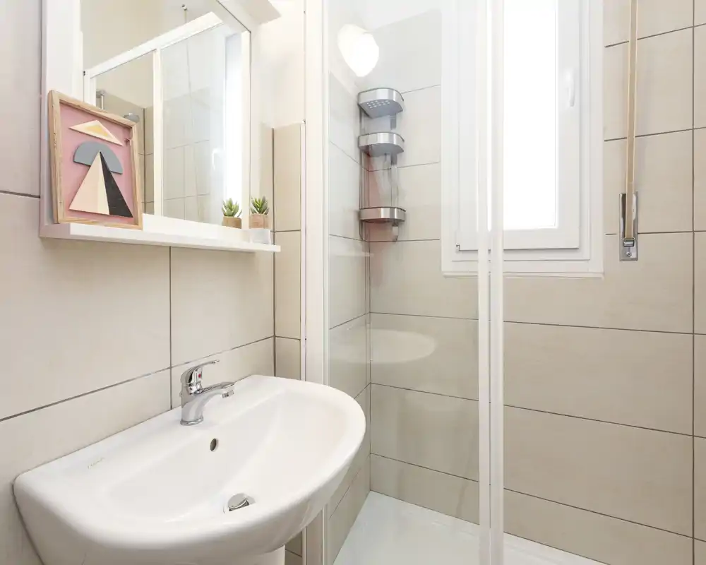 Compact modern bathroom with a white sink and a glass-enclosed shower, tiled walls and a small mirror shelf with decor.