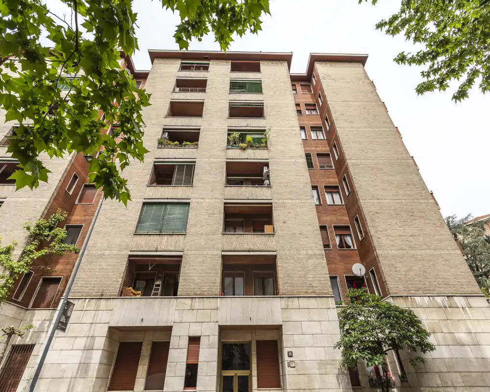 Exterior view of a residential building façade with balconies and the main entrance framed by trees.
