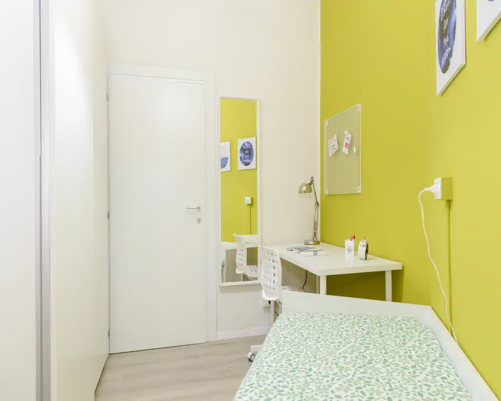 Compact, well-lit bedroom with a single bed, simple desk and mirror — ideal for a student or short-term rental. The bright yellow accent wall and patterned bedding give the space a fresh, modern feel.