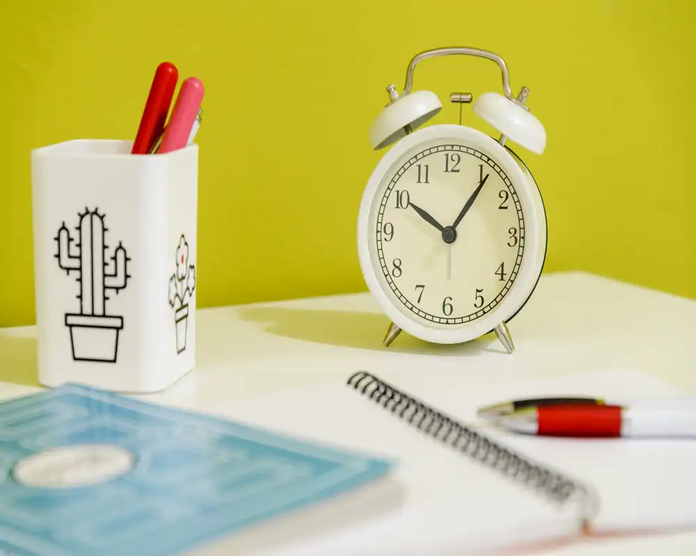 Close-up of a tidy desk corner with a vintage-style alarm clock, pen holder, notebook and pen — a calm workspace detail suitable for listing photos of a study or desk area.