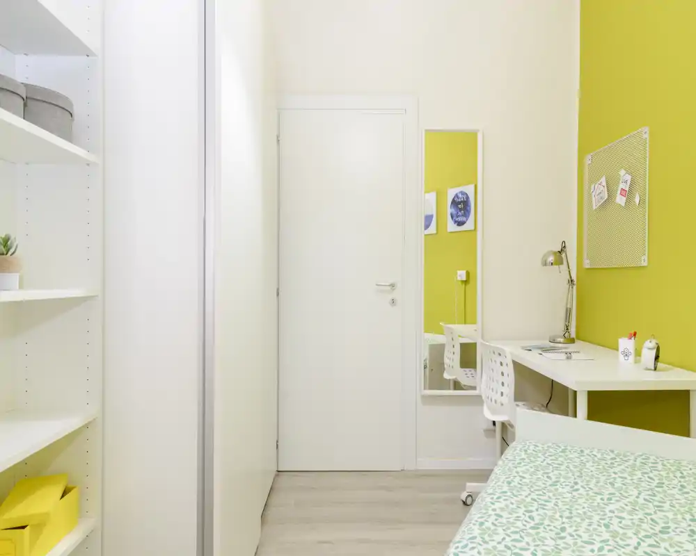 Bright, modern single bedroom with a white desk, shelving and a patterned green bedspread; clean minimalist styling and cheerful chartreuse accent wall.