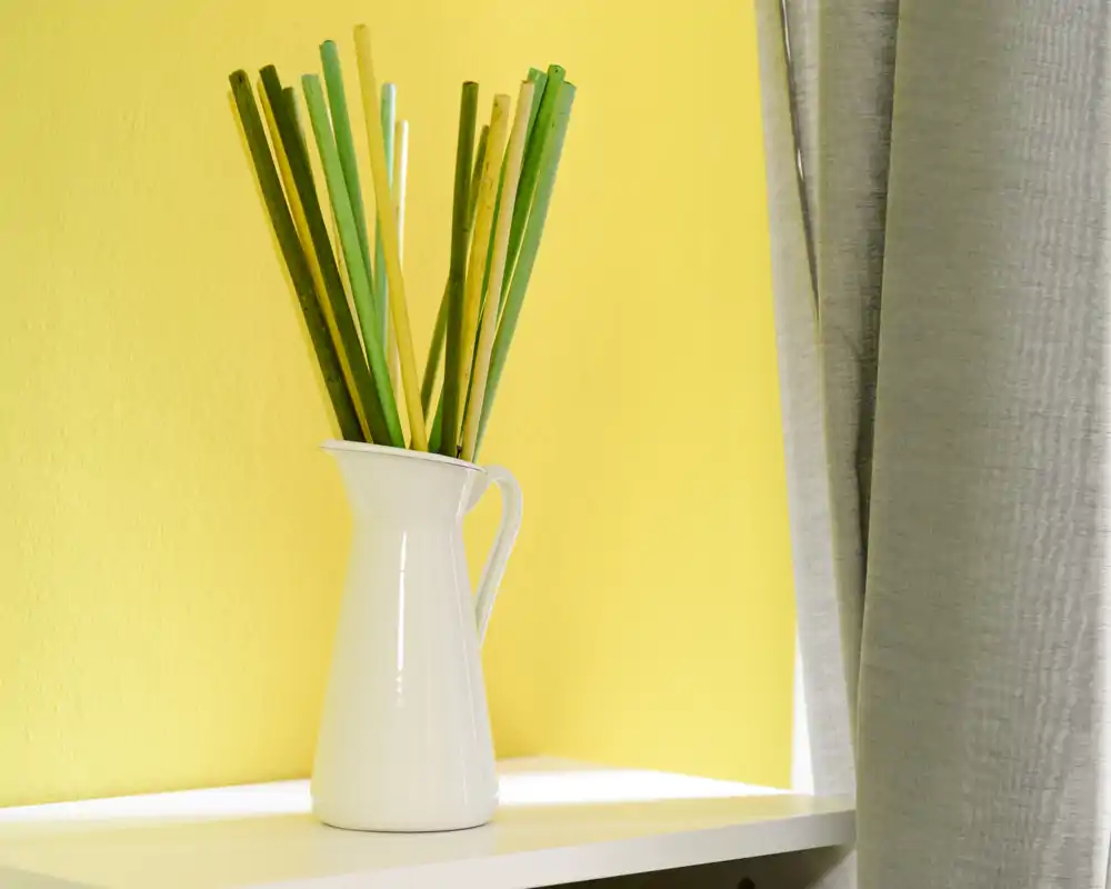 Close-up of a white decorative pitcher used as a vase with tall green and yellow reed-like stems on a white shelf against a bright yellow wall, next to a grey curtain.