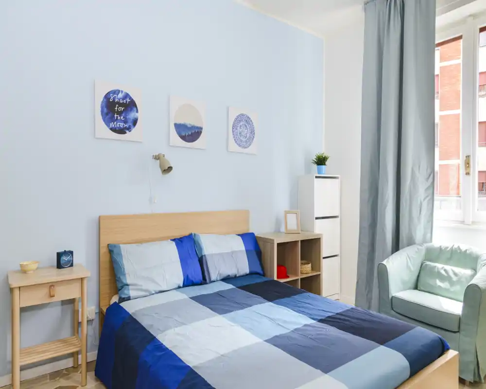 Dormitorio luminoso y bien arreglado con cama doble con ropa de cama a cuadros azules, mesita, estantería y un sillón cómodo junto a la ventana con mucha luz natural.