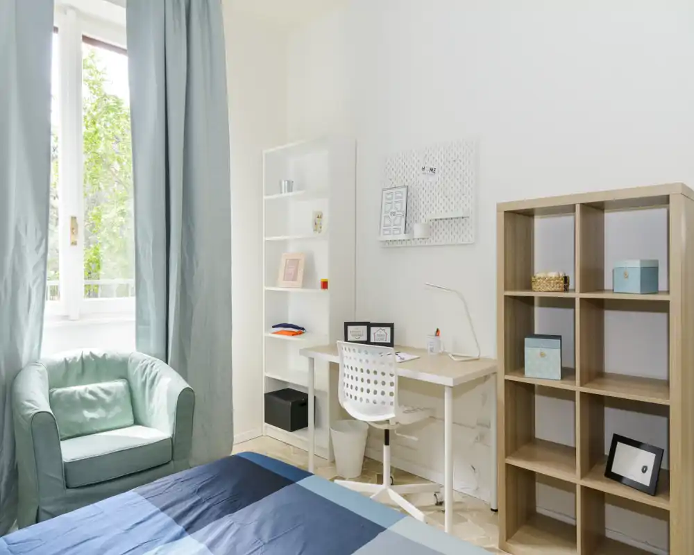 Dormitorio luminoso y ordenado con una cama cómoda en primer plano, escritorio y estanterías—ideal para una persona o estudiante. La luz natural entra por una gran ventana y los textiles azul verdoso crean un ambiente tranquilo y aireado.