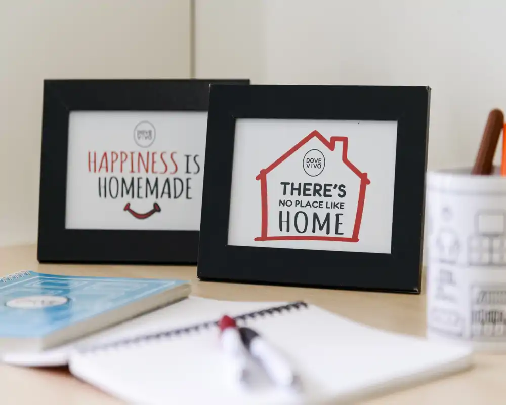 Primer plano de una esquina de escritorio con dos marcos que dicen “Happiness is homemade” y “There’s no place like home”, además de un cuaderno y un bolígrafo desenfocados: un detalle decorativo acogedor para sala o despacho.