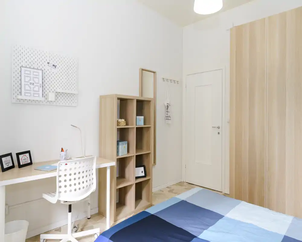 Dormitorio luminoso y minimalista con colcha azul, escritorio sencillo y estantería. El espacio está bien iluminado y ordenado, ideal para una persona o una habitación de invitados.