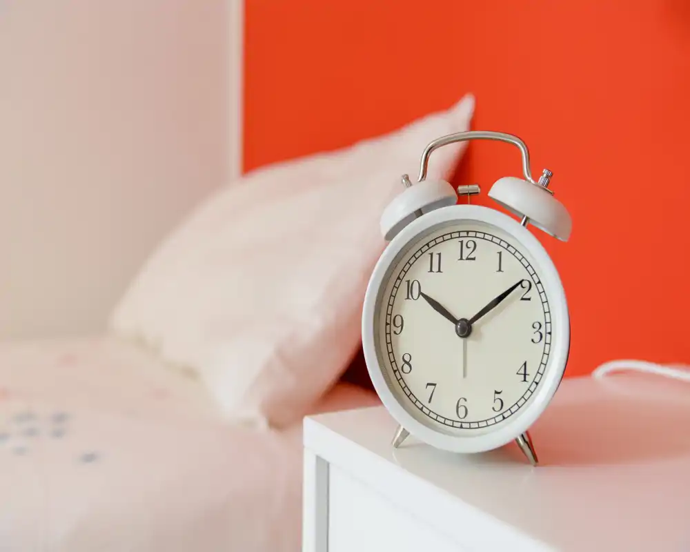 Escena luminosa junto a la cama con un despertador clásico sobre una mesita blanca y almohada parcialmente visible frente a una pared naranja cálida; ideal para destacar detalles de la mesita en un anuncio de dormitorio.