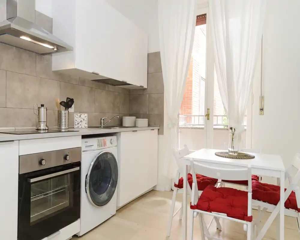 Bright modern kitchen with integrated oven, washing machine and a small dining table by a sunlit door; white cabinets and red seat cushions add a fresh, cozy feel.