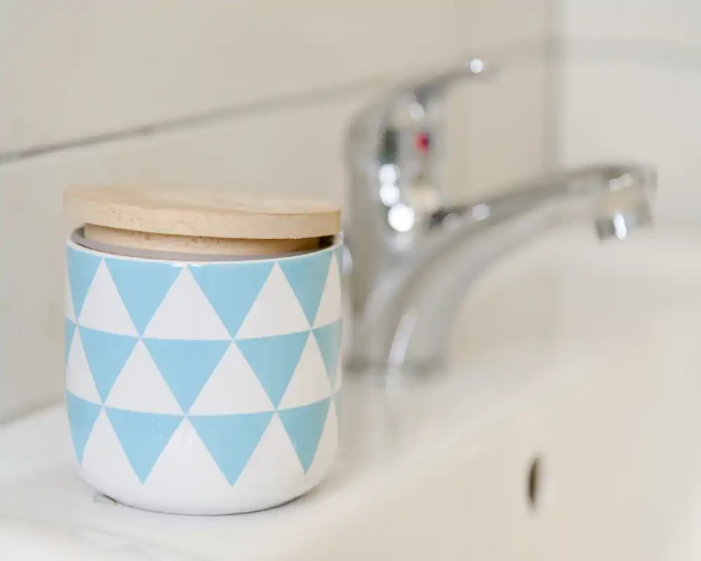 Close-up of a bathroom sink area showing a decorative jar with a wooden lid beside a chrome faucet — tidy and well-lit.