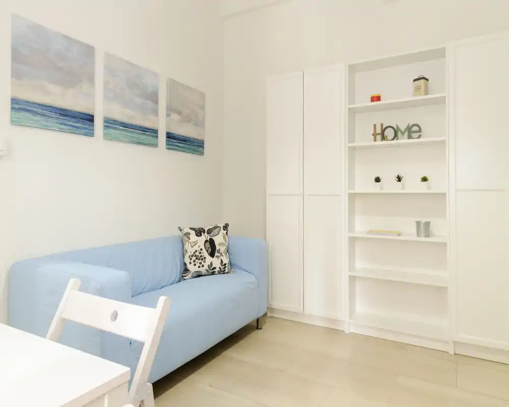 Bright, minimal living area with a light-blue sofa, decorative cushion and shelving; simple coastal artwork and clean white furniture create a fresh, airy space.