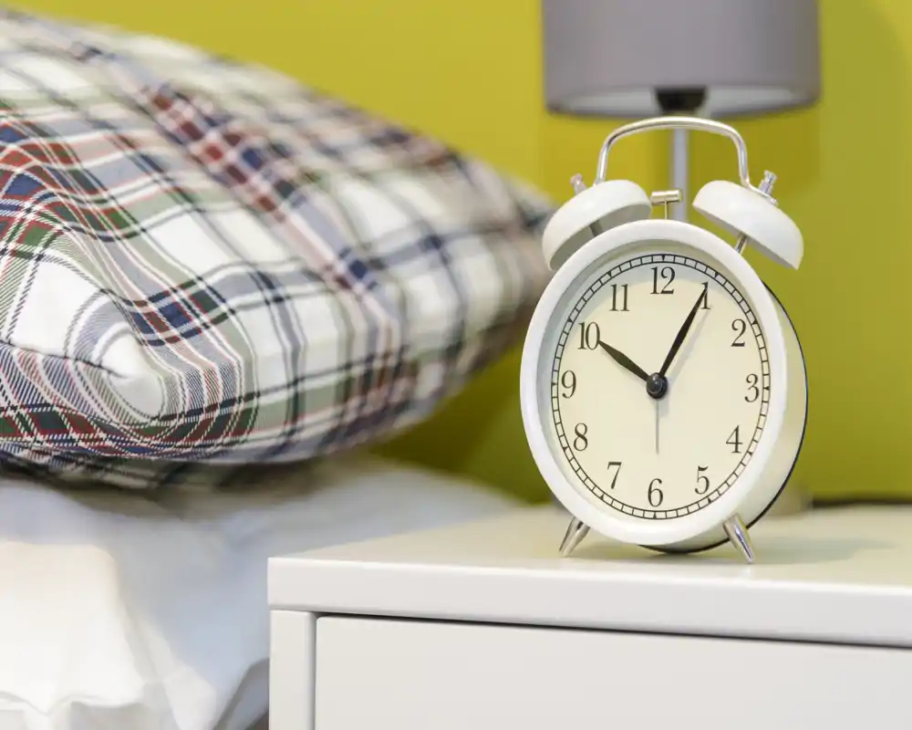 Close-up of a bedroom nightstand with a classic alarm clock, lamp and a plaid pillow visible in the background, suggesting a tidy bedside setting.