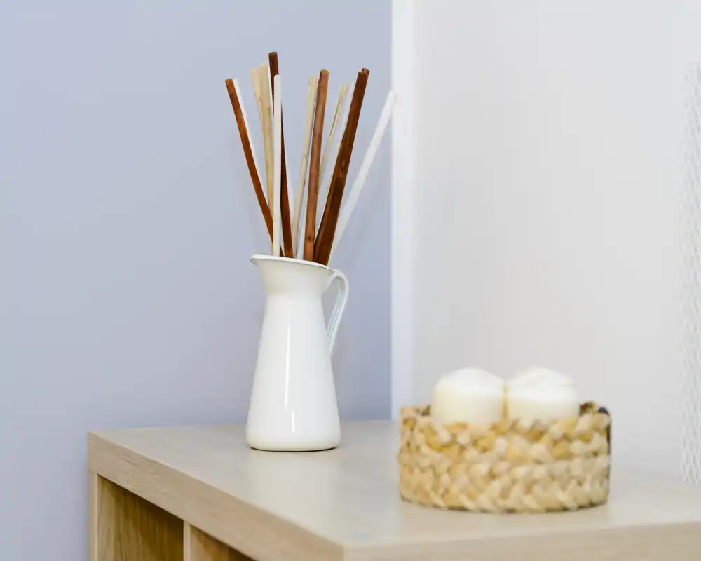 Close-up of decorative accessories on a wooden surface: a white jug with reed sticks and a small woven basket holding candles. The composition highlights styling details suitable for staging or product shots rather than a clearly identifiable room.