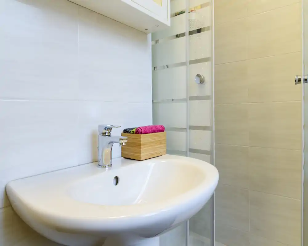 Bright, modern bathroom sink with chrome faucet and a glass shower door. A wooden box with colorful towels adds a neat decorative touch.