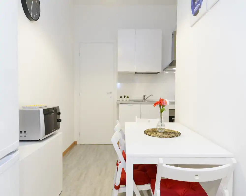 Compact modern kitchenette with a small white dining table and red-cushioned chairs, a countertop sink and upper cabinets; bright, minimal and well-styled for short-stay rentals.