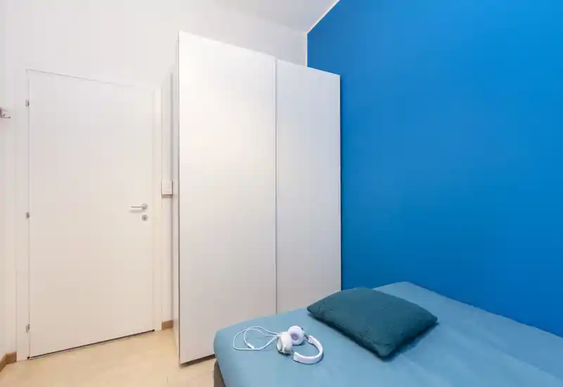 Coin de chambre avec un lit simple contre le mur, literie et coussin bleu sarcelle, une armoire blanche et un mur d'accent bleu vif ; propre, mobilier minimal et lumineux.
