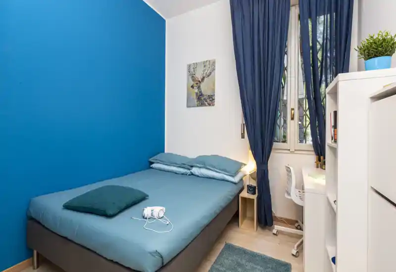 Chambre simple lumineuse et soignée avec mur accent turquoise, lit confortable, lampe de chevet et petit bureau près de la fenêtre — idéale pour un étudiant ou une personne seule.