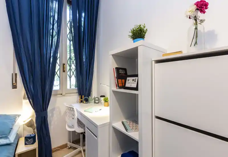 Coin chambre lumineux et bien aménagé avec lit simple, bureau et étagères — idéal pour un étudiant ou un espace de travail. La fenêtre apporte de la lumière naturelle et les rideaux bleus donnent un joli contraste.