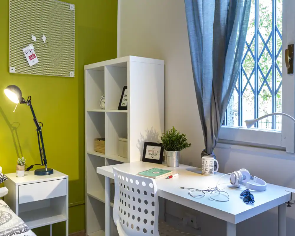 Angolo camera luminoso e moderno con scrivania bianca e libreria accanto a un letto parzialmente visibile. Una finestra con tende blu, lampada da tavolo, pianta e accessori ordinati creano un'area studio funzionale nella zona notte.