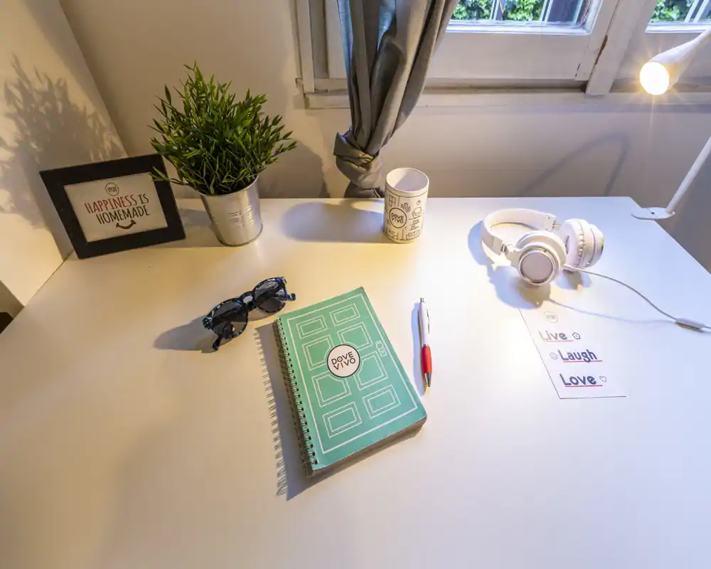 Angolo di studio luminoso e ordinato in camera da letto con scrivania bianca, quaderno, cuffie e pianta decorativa vicino alla finestra; ottima luce naturale e artificiale per lavoro o studio.