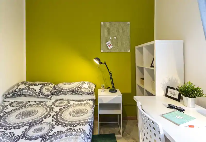 Camera da letto accogliente e ben illuminata con letto matrimoniale a motivo, comodino e scrivania; parete verde a contrasto e scaffalatura ordinata offrono zona notte e lavoro.