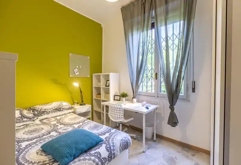 Camera da letto luminosa e ben arredata con un letto matrimoniale a fantasia, lampada da comodino, una scrivania vicino alla finestra e dettagli decorativi che la rendono accogliente.