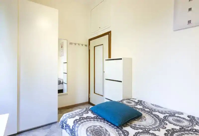 Camera da letto luminosa e ordinata con copriletto decorato e cuscino turchese; mobili bianchi e uno specchio a parete rendono l'ambiente arioso.