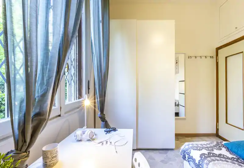 Small bright bedroom corner with a single bed covered in patterned bedding, a white desk by a large window with sheer curtains and a bedside wardrobe.